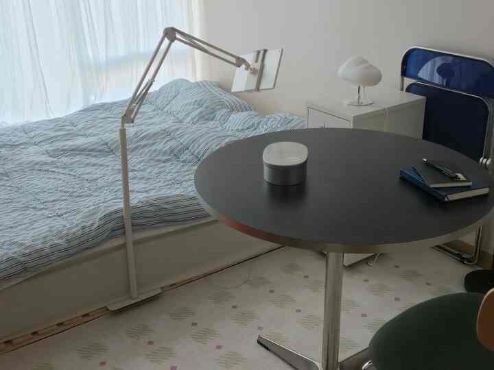 柔らかなパステルカラーのワンルーム、窓際にベッドと丸いダイニングテーブルが配置されたシンプルで居心地の良い空間