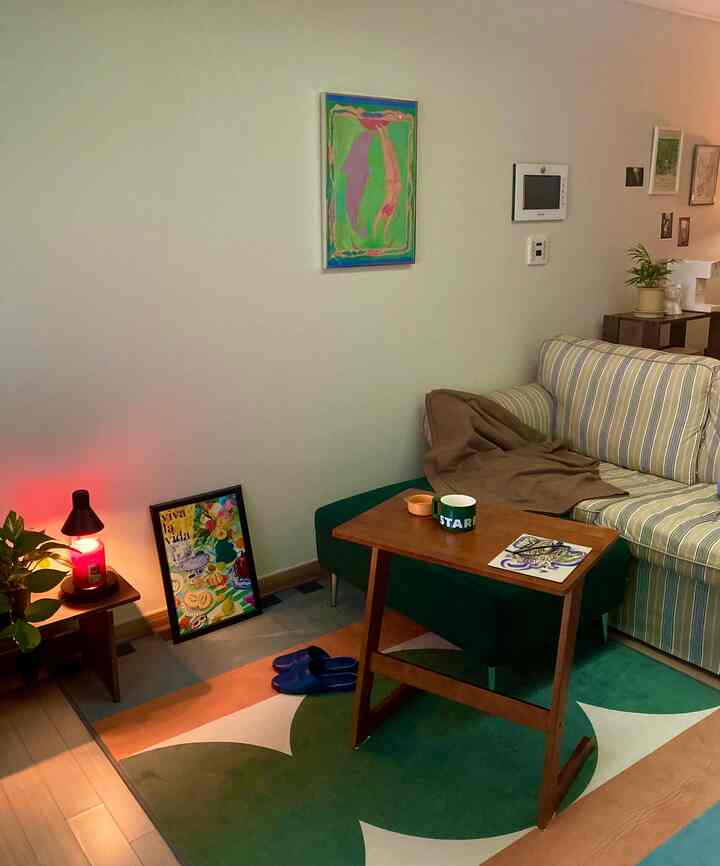 ベージュ色の壁とストライプのソファ、グリーンスツールとカーペットがある居心地の良いリビング、可愛らしいポスターと照明が配置された雰囲気のあるインテリア