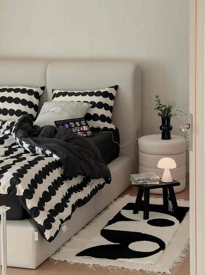 Modern bedroom in white and black tones featuring patterned bedding, rounded stool, and a table lamp, creating a cozy atmosphere