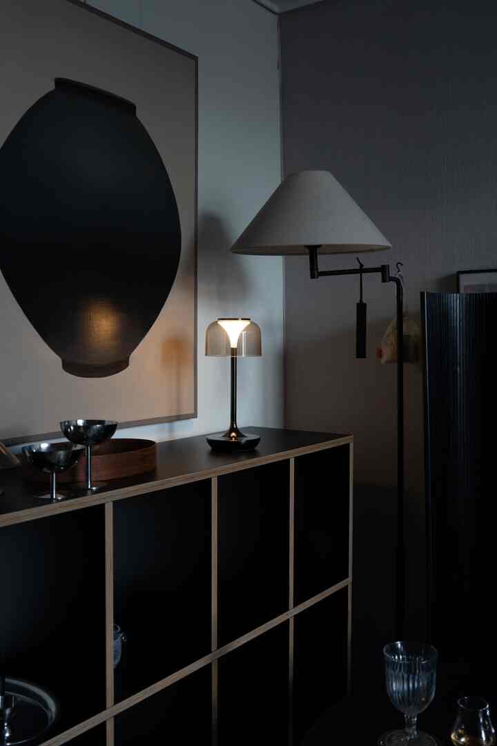 暗めのトーンのリビングスペース、ブラックチェストの上に透明のテーブルランプとベージュのフロアランプが配置されたシンプルでモダンなインテリア