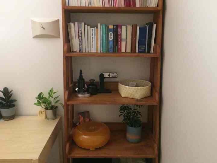 白い壁と茶色い木製の本棚がある小さな寝室、書籍やオブジェ、植物が調和したナチュラルなインテリア