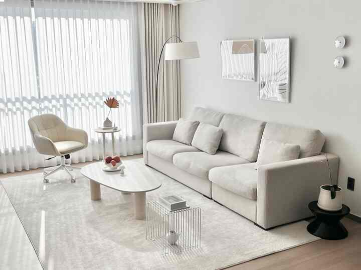 淡いベージュとホワイトのトーンのリビング、クリーム色のソファと回転式アームチェア、丸いサイドテーブルとラグが配置されたモダンで清潔感のあるインテリア