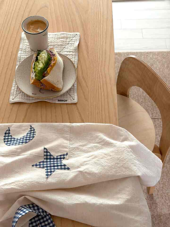 ナチュラルなウッドトーンのダイニングテーブルと椅子、星と月のパッチが付いたエプロンが配置された雰囲気のあるおうちカフェ空間