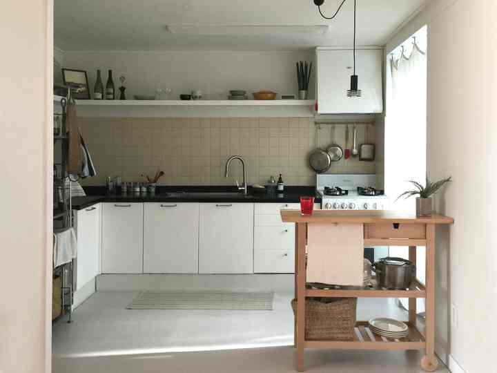 白とベージュのトーンのミニマルキッチン、木製のキッチンワゴンと壁棚が配置されたナチュラルでシンプルなインテリア