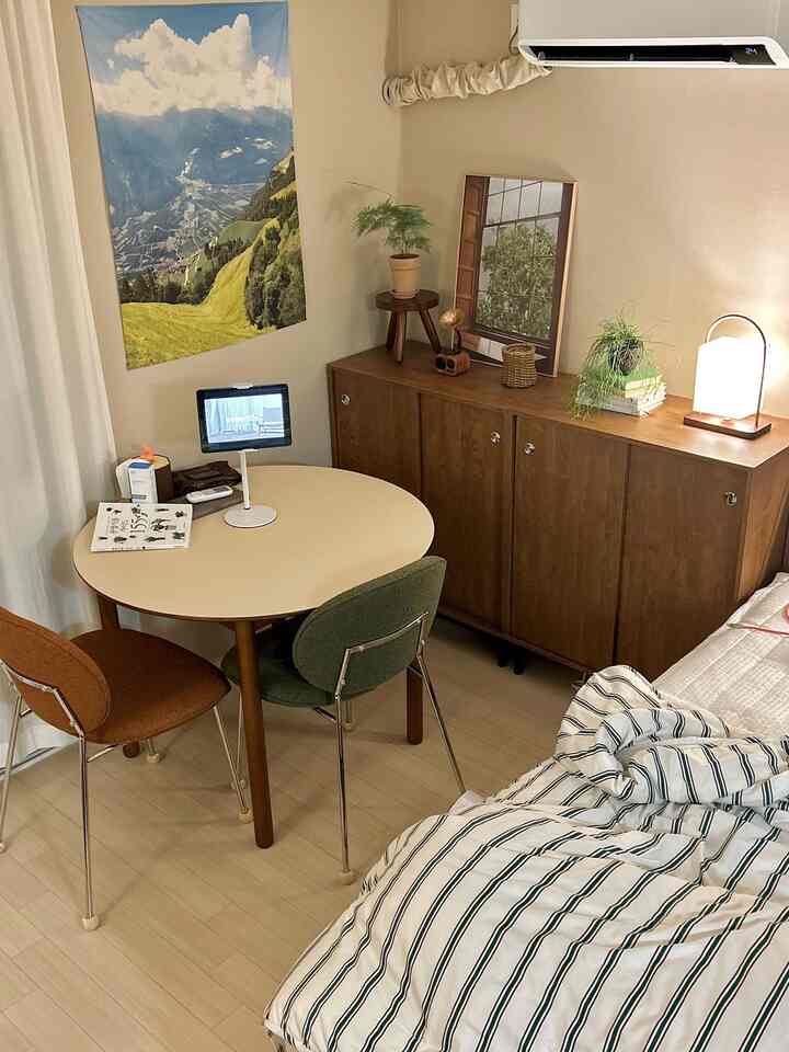 ベージュとブラウンのナチュラルトーンのワンルーム寝室、丸テーブルとブラウン・グリーンのダイニングチェアが配置された雰囲気のある空間