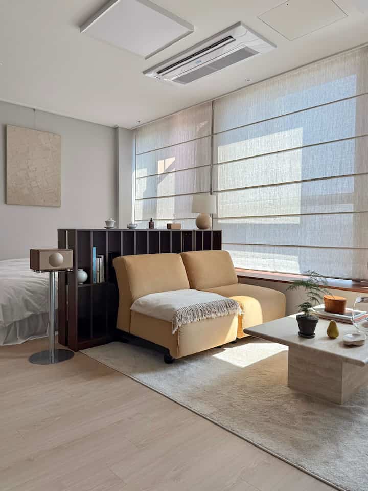 ベージュ調のリビング、リネンのローマンシェードとベージュのソファが配置された落ち着いた雰囲気の空間