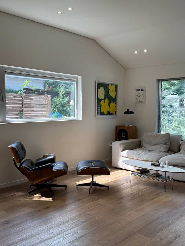 明るいホワイトの壁と木目調の床が特徴のリビング、イームズラウンジチェアとベージュのソファ、壁時計とアート作品が調和した雰囲気のあるインテリア