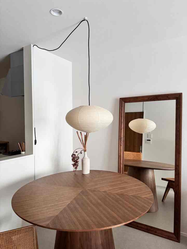 白いトーンのキッチンに茶色の丸い木製ダイニングテーブル、大きな木枠の鏡とペンダントライトが配置されたモダンで自然な空間