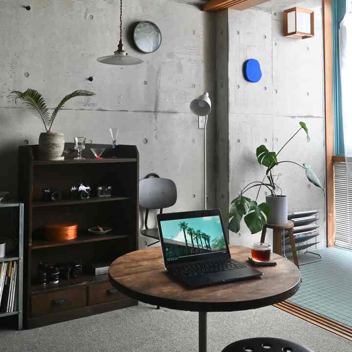 コンクリートの壁と丸い木製テーブルがあるリビング、ノートパソコンと植物が配置されたモダンな雰囲気の空間