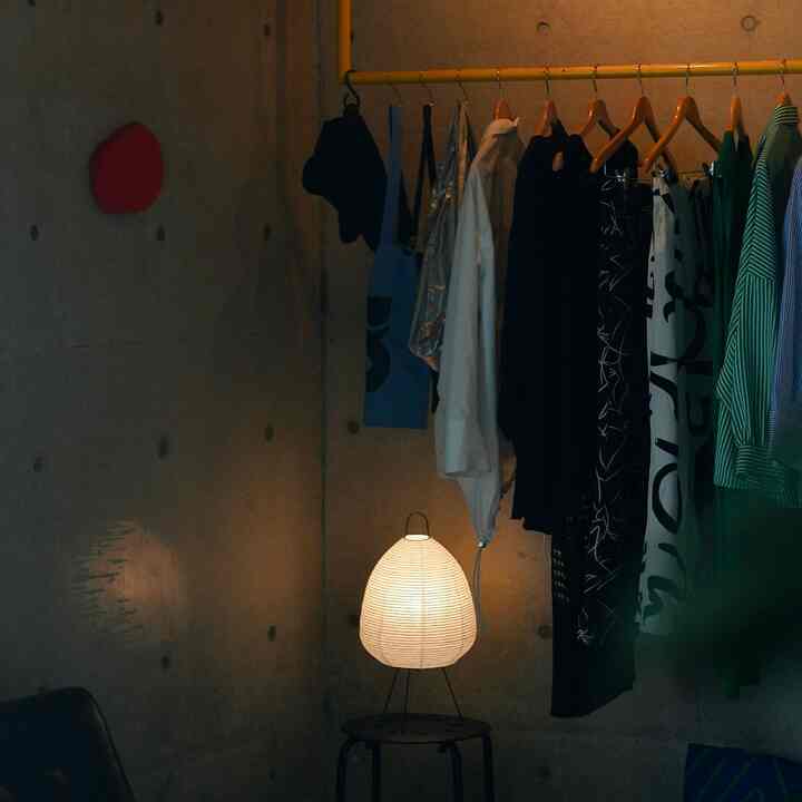 黄色のハンガーラックと木製ハンガーが置かれたインダストリアル風の空間、柔らかな光の和風紙のフロアランプが配置されている