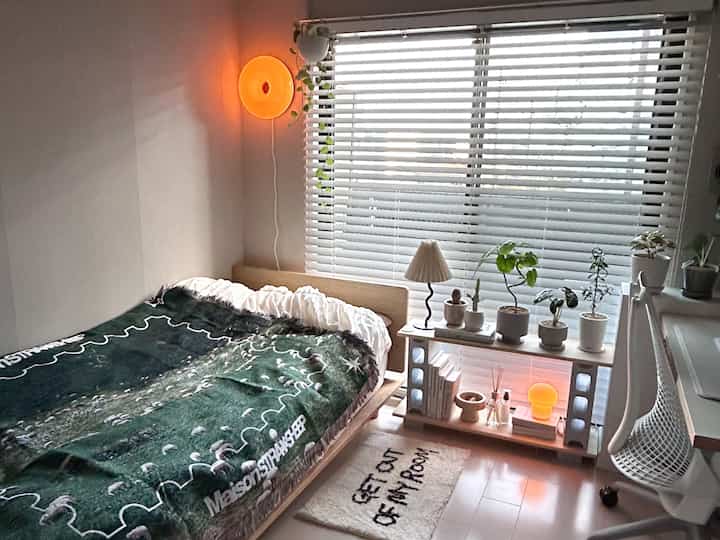 ベージュ色調の小さな寝室、窓際に植物と照明が配置された自然な木製ベッドフレームのある雰囲気のあるインテリア