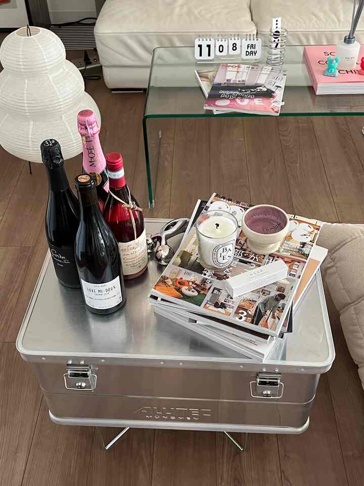 シルバー調のリビング、アルミコンテナのサイドテーブルとガラスのコーヒーテーブルがあり、ワインボトルと本が置かれたシンプルな空間