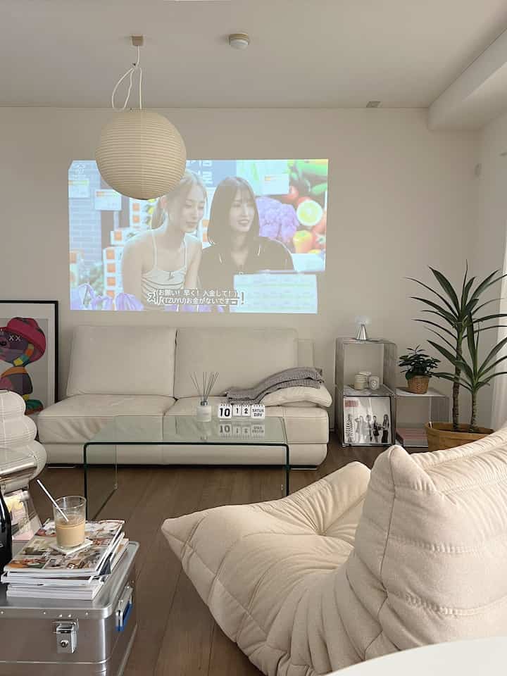 白とベージュの落ち着いたトーンのリビング、プロジェクターの映像、ソファ、ガラスのコーヒーテーブル、植物が配置された雰囲気のあるインテリア