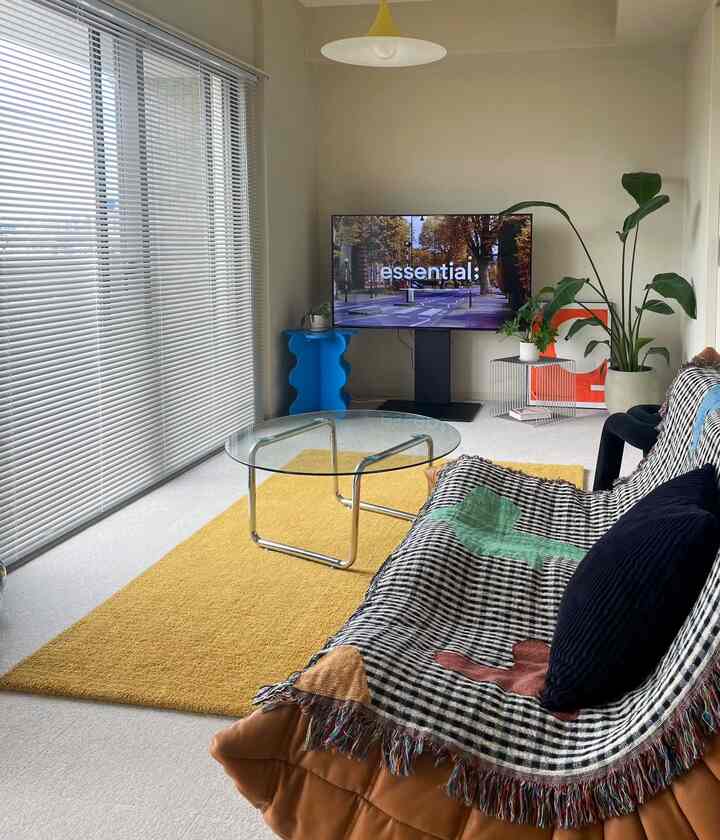 白い壁と天井の6畳リビング、黄色のラグと透明なコーヒーテーブル、茶色の2人掛けソファ、大きな植物が配置されたモダンな空間