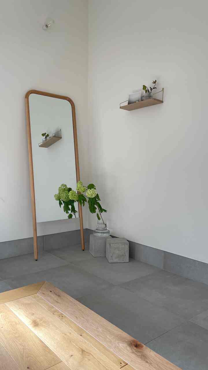 白い壁とグレーのタイル床の玄関、木製フレームの鏡と小さな棚が配置されたミニマルで自然な雰囲気のある空間