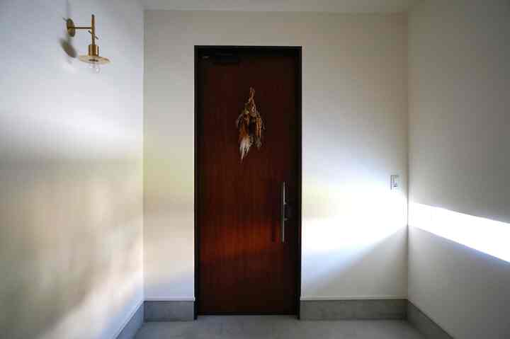 白い壁とコンクリート床の玄関、中央に茶色い木製のドア、左壁にブラス製の壁照明が配されたシンプルで落ち着いた雰囲気の空間