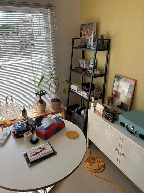 白と黒を基調とした7畳の狭い部屋、丸いダイニングテーブルと金属棚、キャビネット、植物が配置されたナチュラルで落ち着いた雰囲気の空間