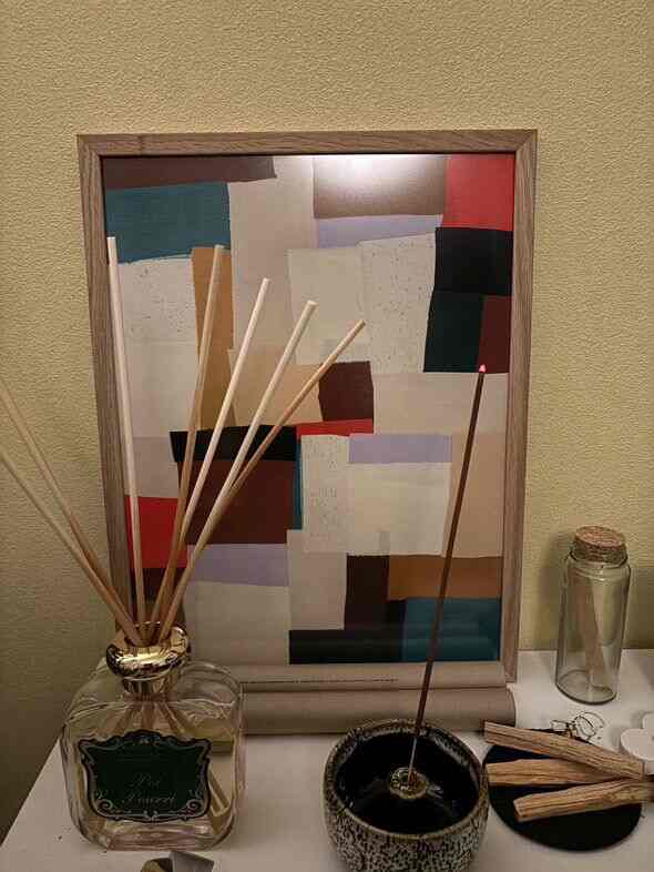 ベージュの壁を背景に、小さなテーブルの上にディフューザー、香立て、木製の香、抽象画が配置された自然な雰囲気のインテリア小物の空間