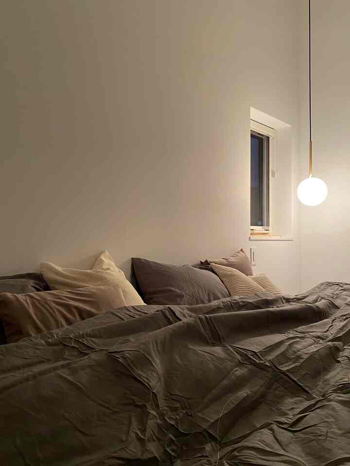 グレーとベージュの色調の寝室、ベッドに様々なクッションと寝具が配置され、右側にペンダントライトが灯る雰囲気のある空間