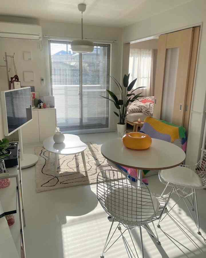 白を基調としたリビングと隣接する寝室、白い家具とブラインド、大型観葉植物が配置された明るく清潔感のある空間