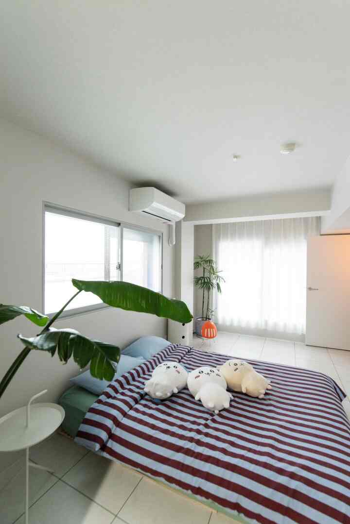 明るいナチュラルな白色の寝室、中央にストライプのベッドカバーが敷かれたベッドと緑の植物が配置された空間