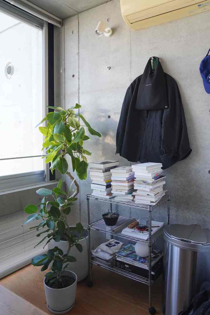 ナチュラルカラーとコンクリート壁が調和する狭い部屋のコーナー、金属製の棚に積まれた本、掛けられた服と置かれた植物が配置された空間