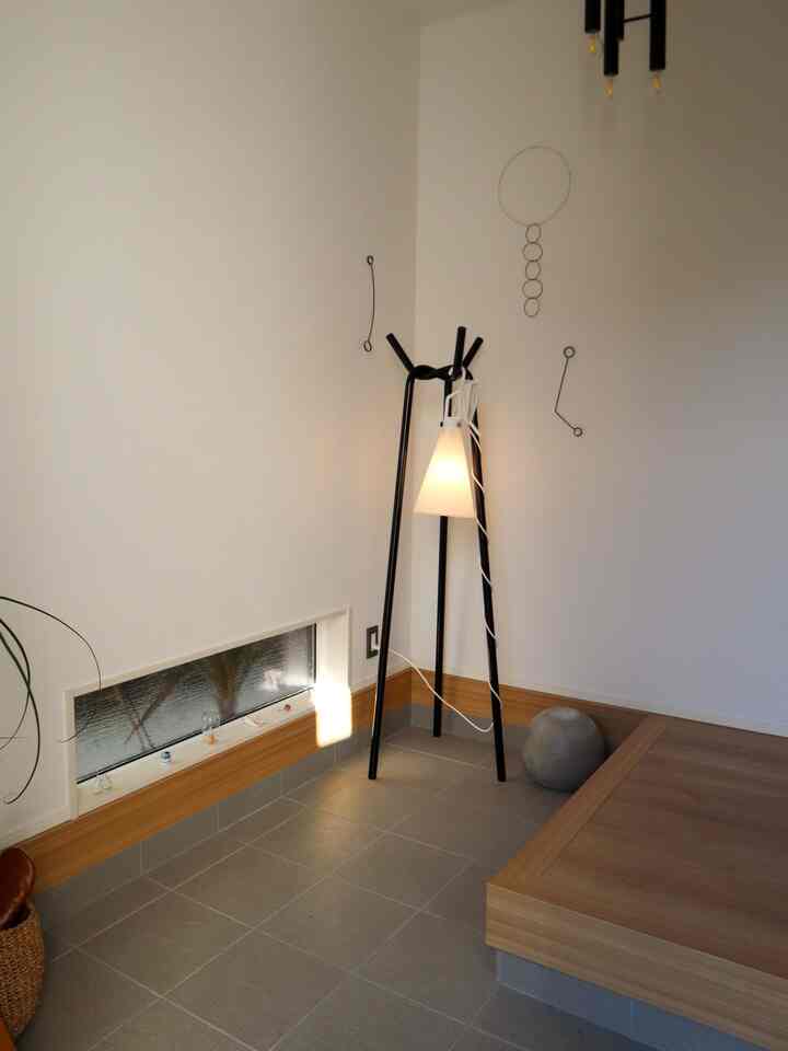 白い壁とグレーのタイル床のL字型玄関、黒のコートハンガーと洗練されたフロアランプが配置されたナチュラルモダンな空間