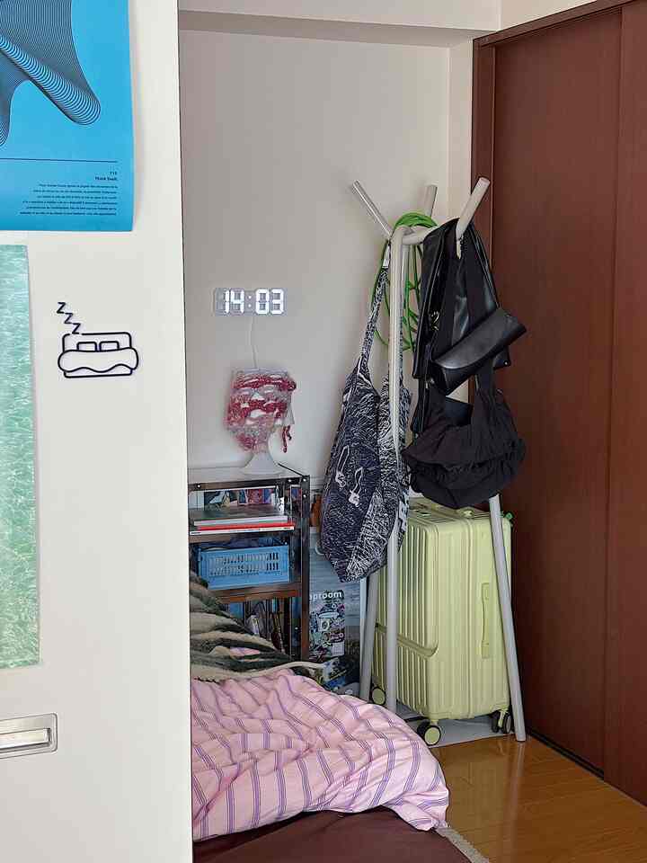 白い壁と薄い木目の床の狭い寝室、ピンクのストライプの寝具、バッグが掛かったコートラック、キャリーバッグが置かれた落ち着いた個人空間