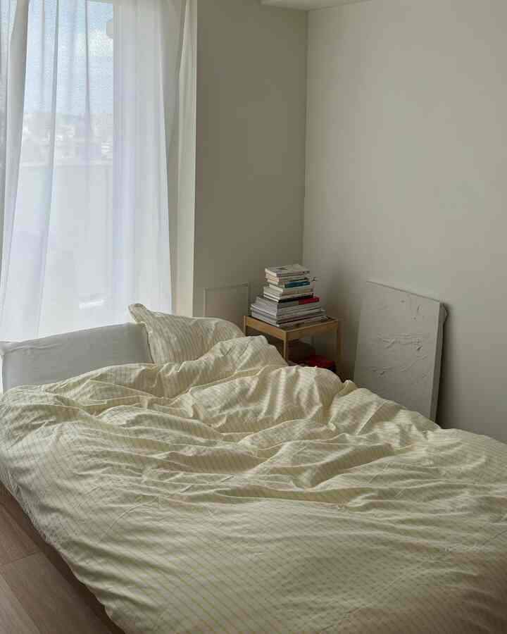 明るい黄色のストライプ柄のベッドカバーと枕が配置された清潔感のある寝室、横に本が積まれた小さな木製サイドテーブルあり