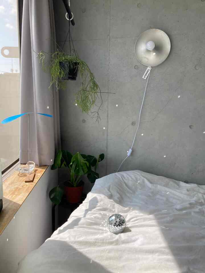 明るい光に包まれたミッドセンチュリーモダンスタイルの寝室、白い寝具とシルバーのブラケットライト、窓際にミラーボールと植物が配置された印象的な空間