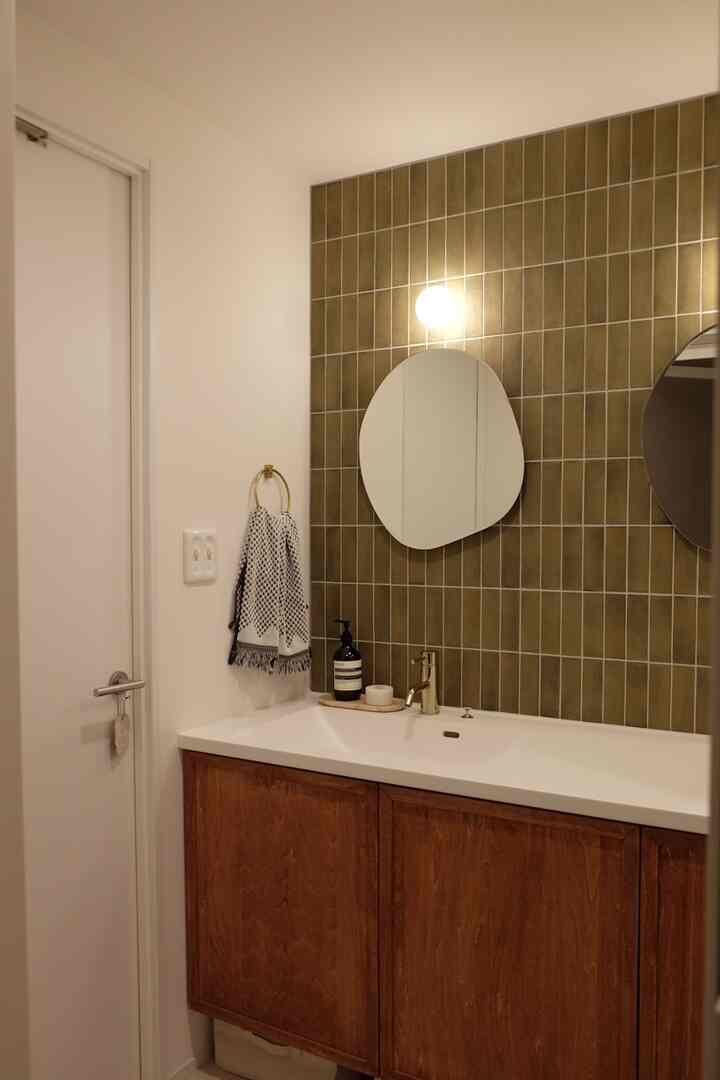 白い壁と木目調のキャビネットが配置されたシンプルモダンな洗面スペース、オリーブグリーンのタイルとゴールドのアクセサリーがポイントの落ち着いた雰囲気のインテリア