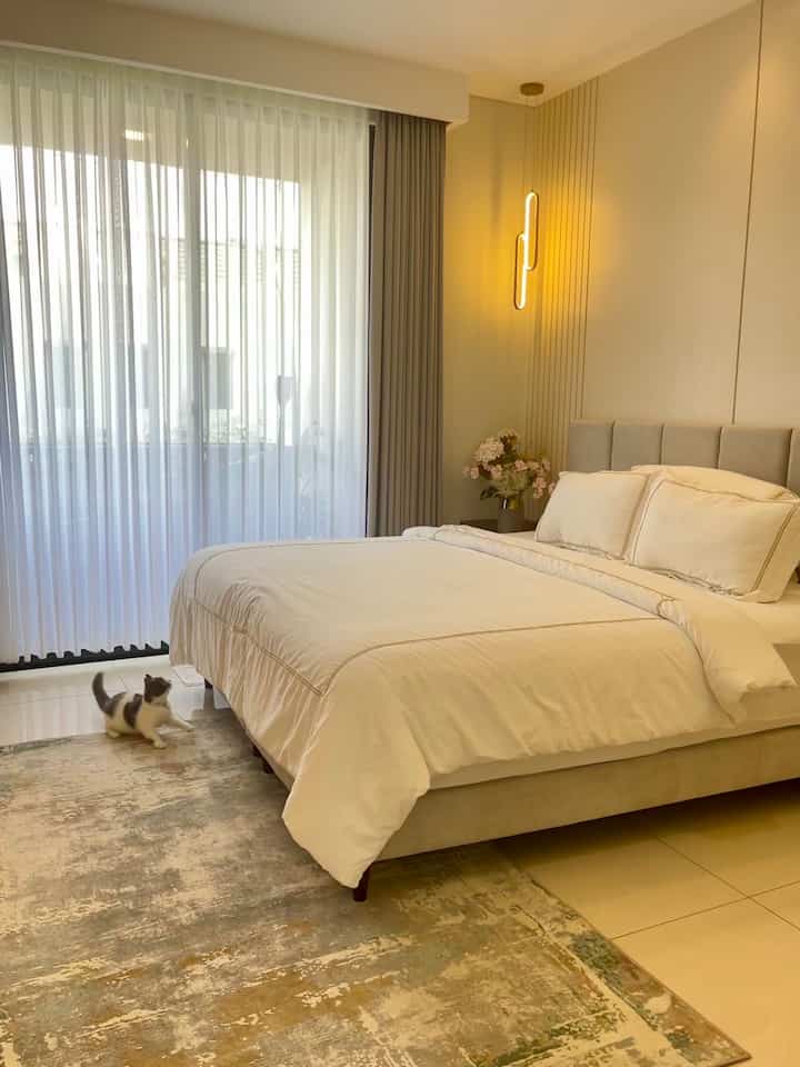 White and beige toned bedroom featuring a large bed, carpet, and pendant light creating a cozy atmosphere with a cat on the floor