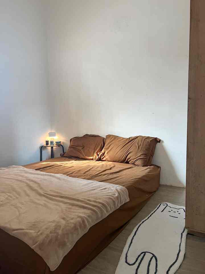 Natural brown and beige toned bedroom featuring a bed with simple bedding, a small side table, and warm table lamp creating a cozy atmosphere