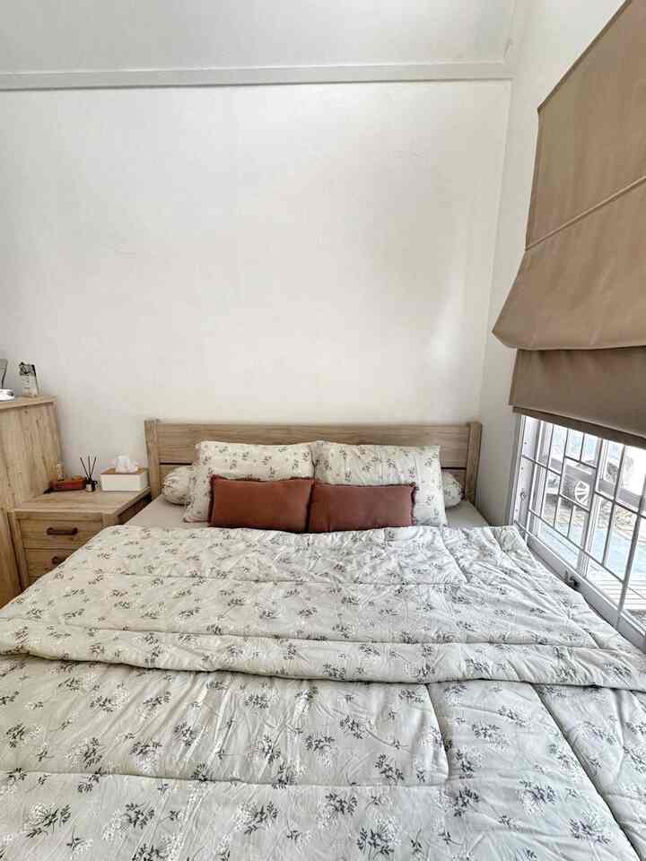 A small bedroom with white walls and wood tone furniture, featuring natural bedding and beige curtains creating a cozy atmosphere
