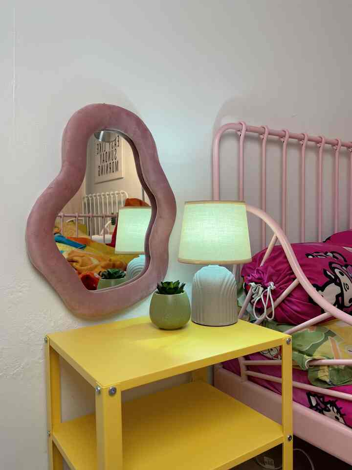 A cozy kids' bedroom with pink and yellow tones featuring a pink velvet framed mirror and yellow nightstand against a white wall