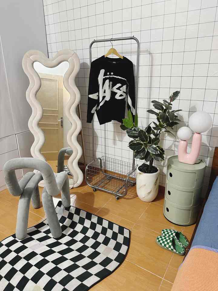Modern bedroom with white tiled walls and wood-tone flooring, featuring a wavy mirror, unique stool, and green side table