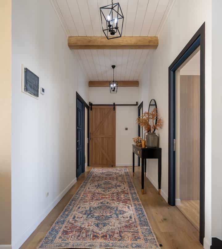 White walled long and narrow hallway with wood tone beams, farmhouse-style console table, and patterned carpet runner