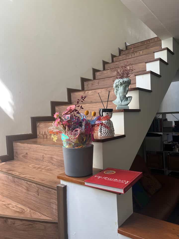 We decorated the stairwell area with various vases and piles of books to make it more lively. The walls of the stairs were broken down and given wooden accents so that more sunlight could enter the living room. #stairwaydecor #stairwaystyling #livingroomstyling #midcenturymodern #rumahcluster