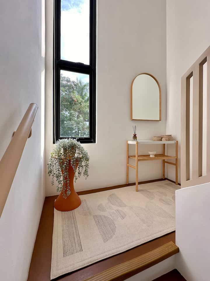 Natural-toned small stair landing featuring an arched mirror, wooden side table, terracotta plant pot, and patterned rug