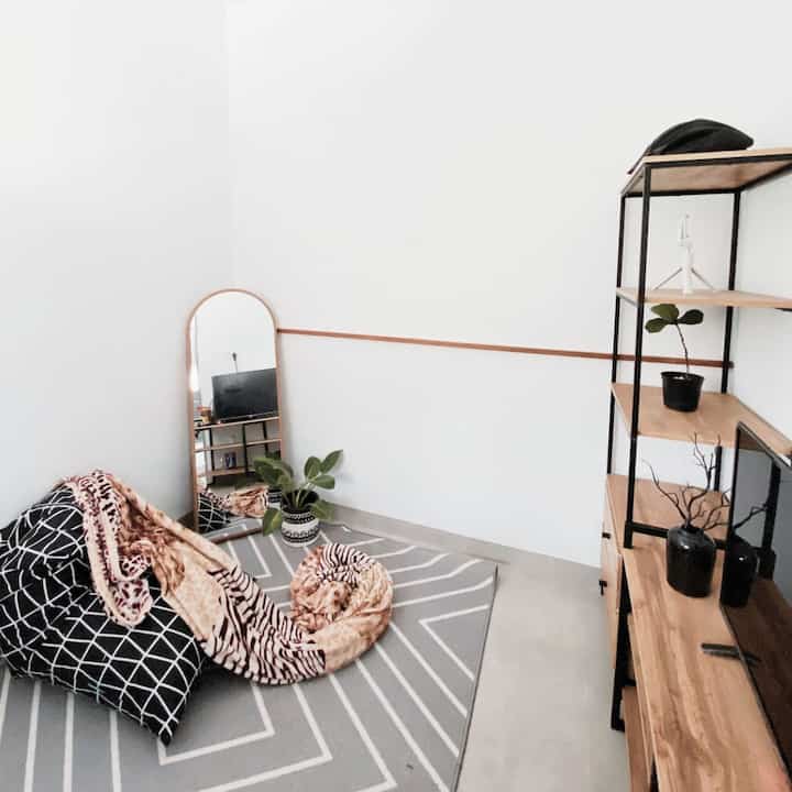 A compact living room in white and gray tones featuring a wooden shelf, bean bag, and full-length mirror with a simple natural vibe