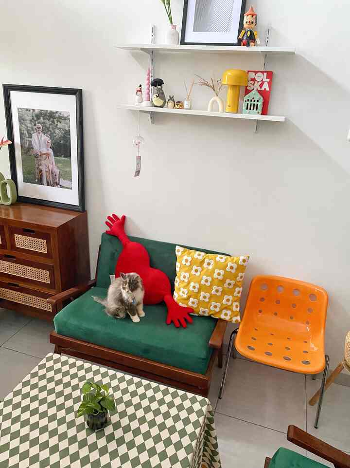 Natural tone living room with white walls featuring wall art and shelves, green sofa with a cat and orange chair creating a cozy atmosphere