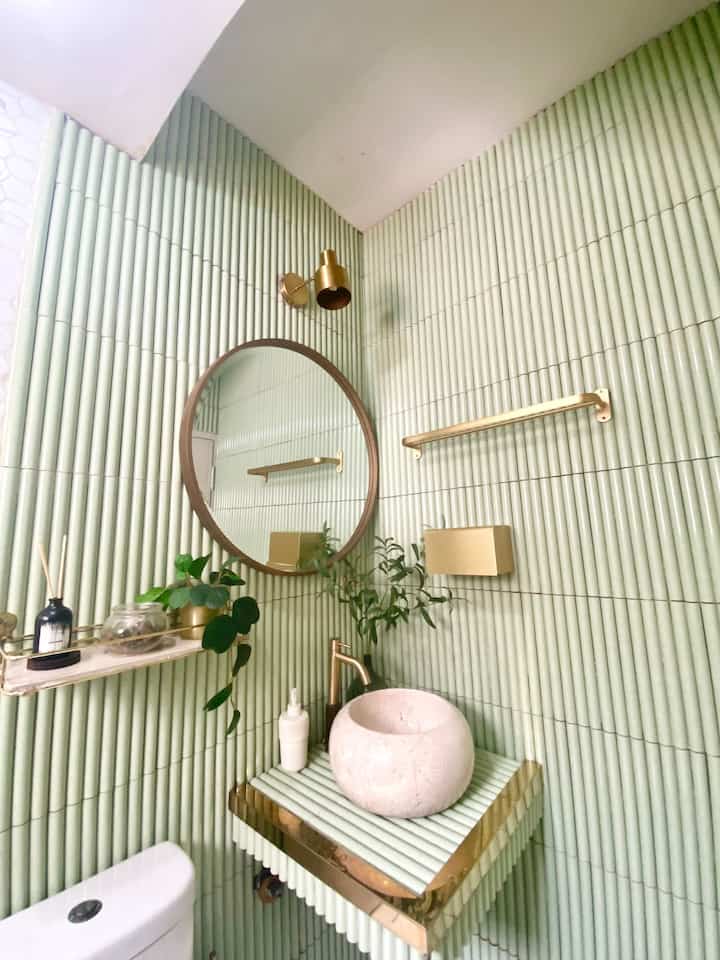Small restroom with dominant green tiles and gold fixtures, featuring a round mirror and natural plants for a clean aesthetic