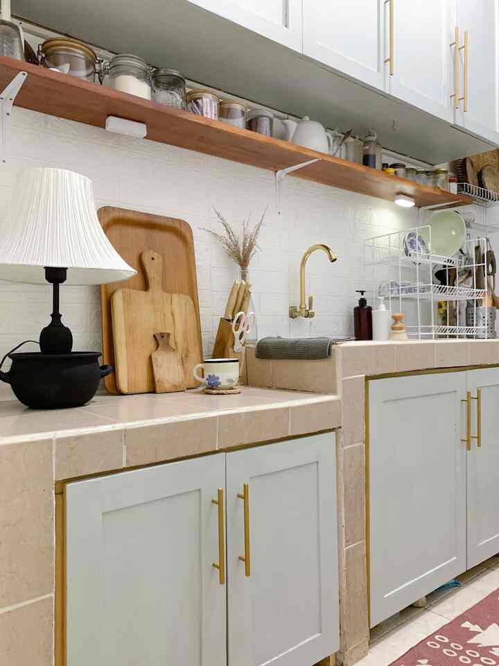Kitchen space with white tiled walls, wood tone shelf, gold handles, and a table lamp creating cozy atmosphere