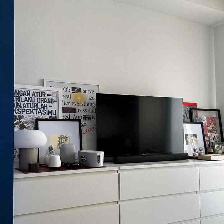 A white-toned studio apartment living room featuring a TV stand with posters and diffusers, showcasing a modern and clean space