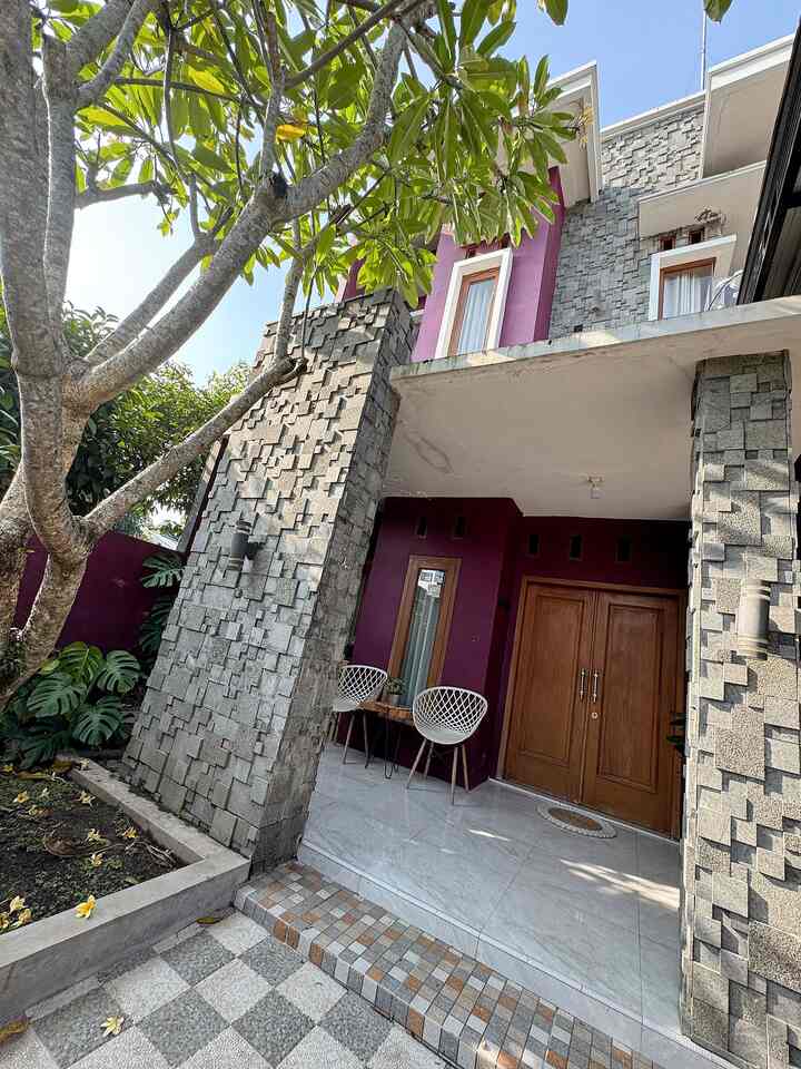 Purple-toned exterior entrance with green plants, wooden door, and textured gray stone walls under natural daylight