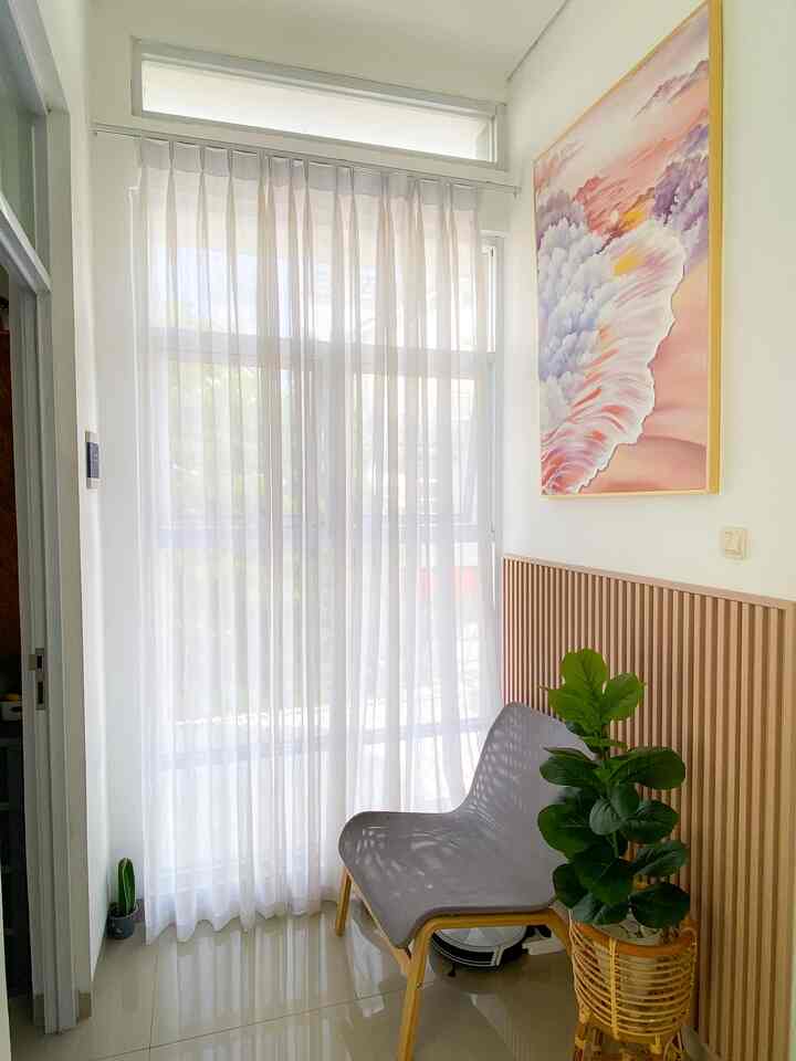 Bright white and natural wood-toned small second-floor relaxation space featuring a single gray armchair, large green plant, and a pink-orange beach-themed framed painting, creating a cozy and calm interior