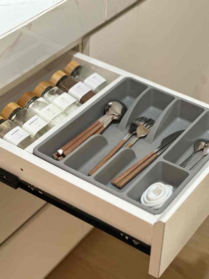 Transparent glass spice bottles and wooden-handled cutlery neatly organized in kitchen drawer, featuring wood and gray tones