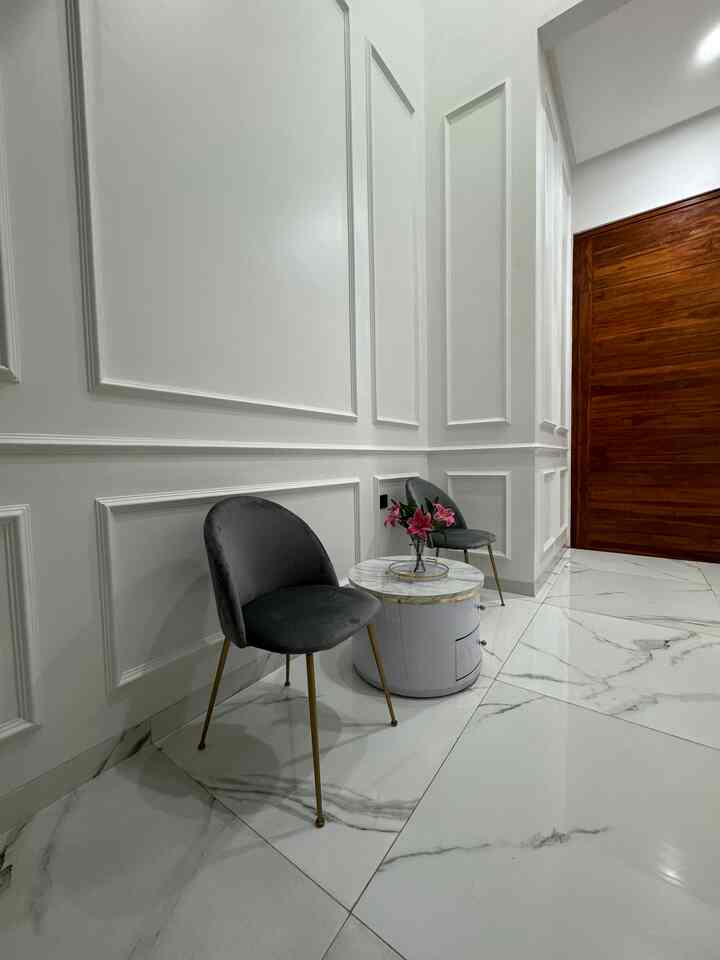 A luxurious entrance area featuring white walls and marble flooring, with two gray chairs with gold legs and a round side table arranged elegantly