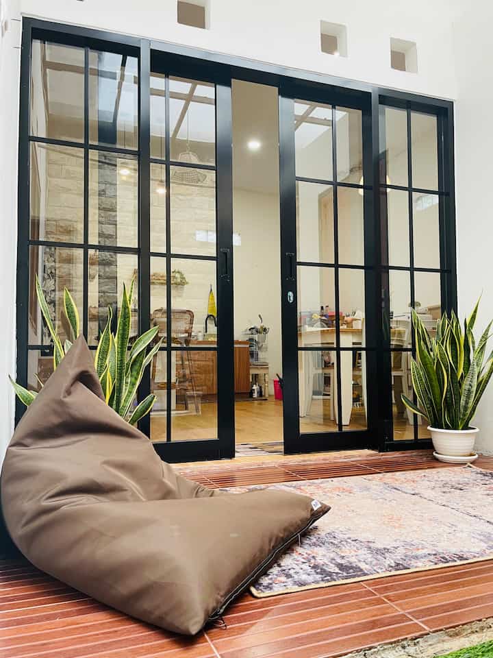 Natural outdoor space featuring a brown bean bag and carpet in front of dark-framed sliding glass doors, flanked by two large plants on both sides