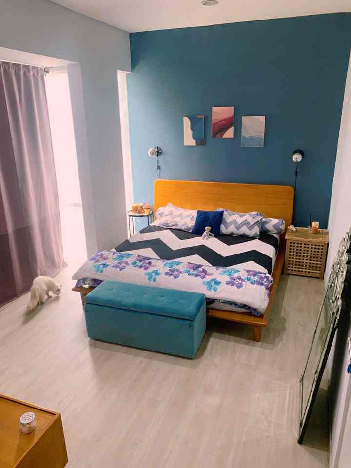 Cozy blue-toned bedroom featuring a wooden bed with patterned bedding, wall decor, and soft curtains in a simple setting